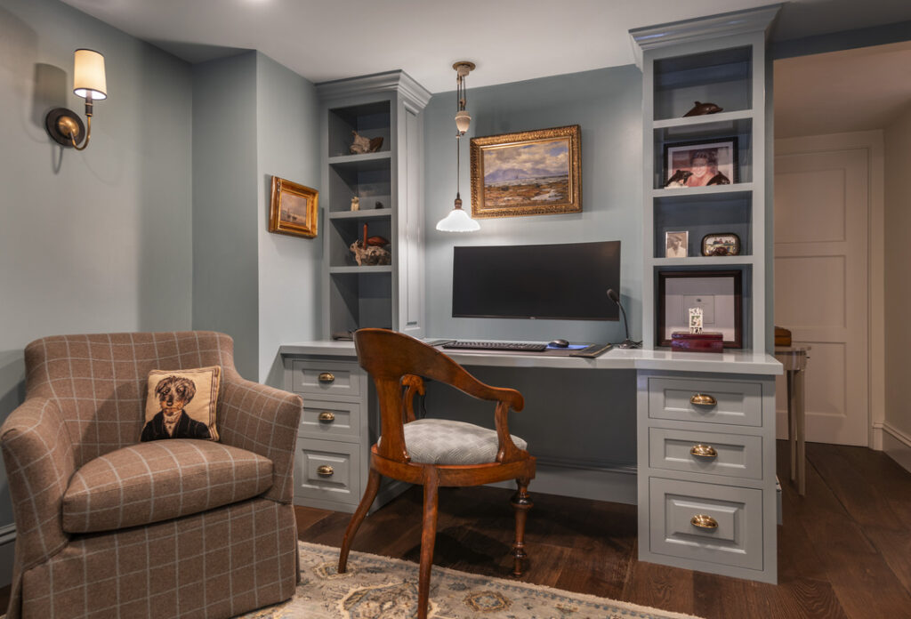 A home office by MBA Builders features built-in shelves, a large monitor, and a wooden chair. An armchair with a dog cushion adds comfort. Light blue walls create a serene atmosphere, while the pendant light above the desk completes this inspiring portfolio space.