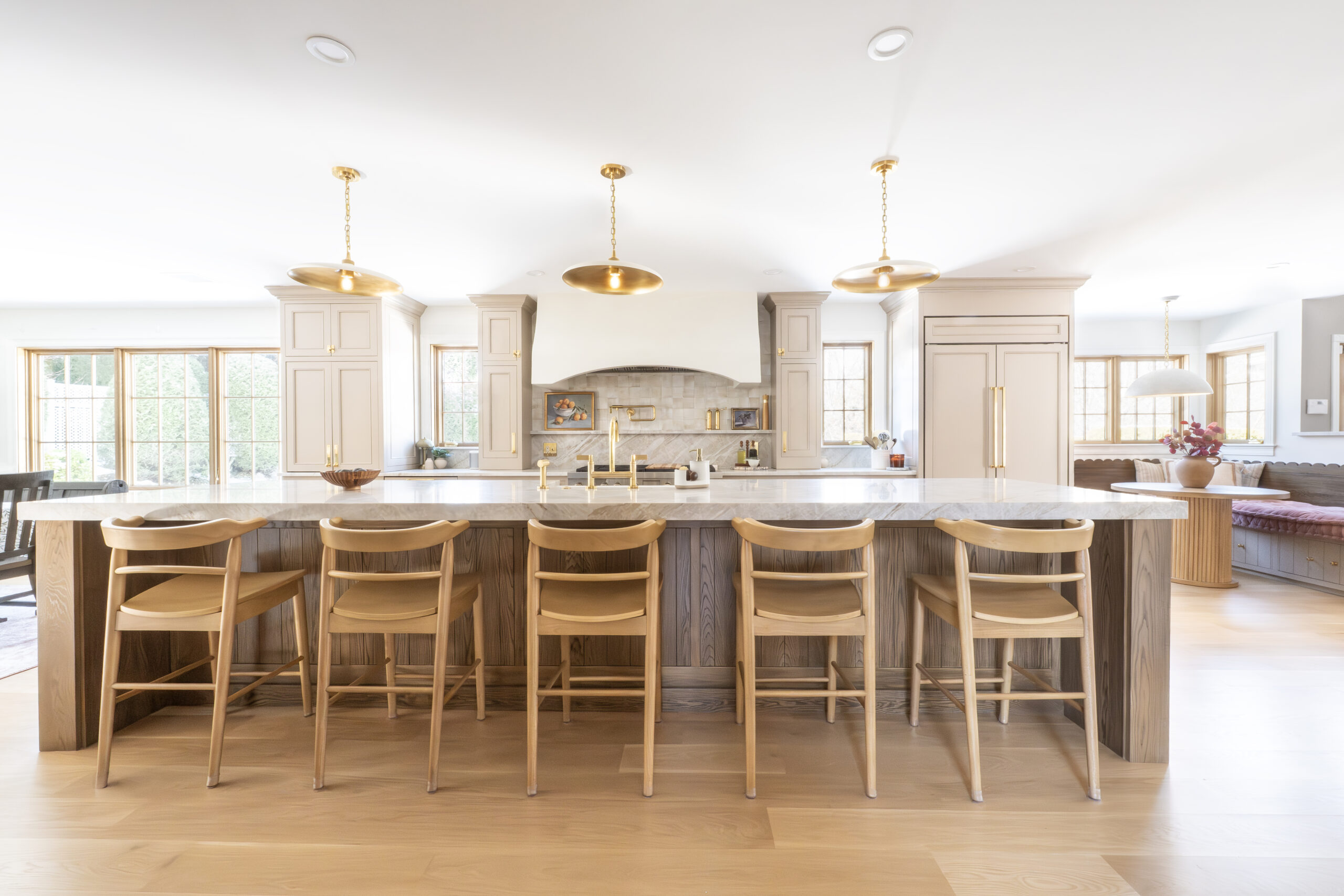 Spacious kitchen with a large island featuring six wooden chairs. Modern lighting hangs above, and the room has light wood cabinetry and large windows allowing ample natural light. example of dining room and kitchen renovation in Reading MA by MBA Builders with custom build finishing and modern kitchen aesthetic