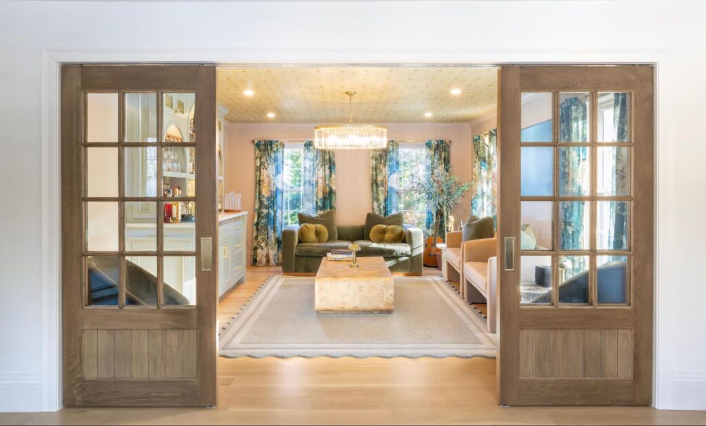 A living room by a custom home builder Lexington, featuring sliding wooden doors, two green armchairs, a beige sofa, a patterned ottoman, and large windows with floral curtains. A living room by a custom home builder Lexington, featuring sliding wooden doors, two green armchairs, a beige sofa, a patterned ottoman, and large windows with floral curtains.