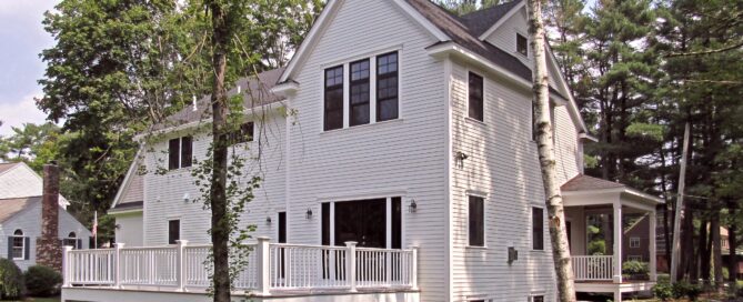 Two-story white house with black-framed windows, elevated deck with white railing, surrounded by green grass and tall trees.