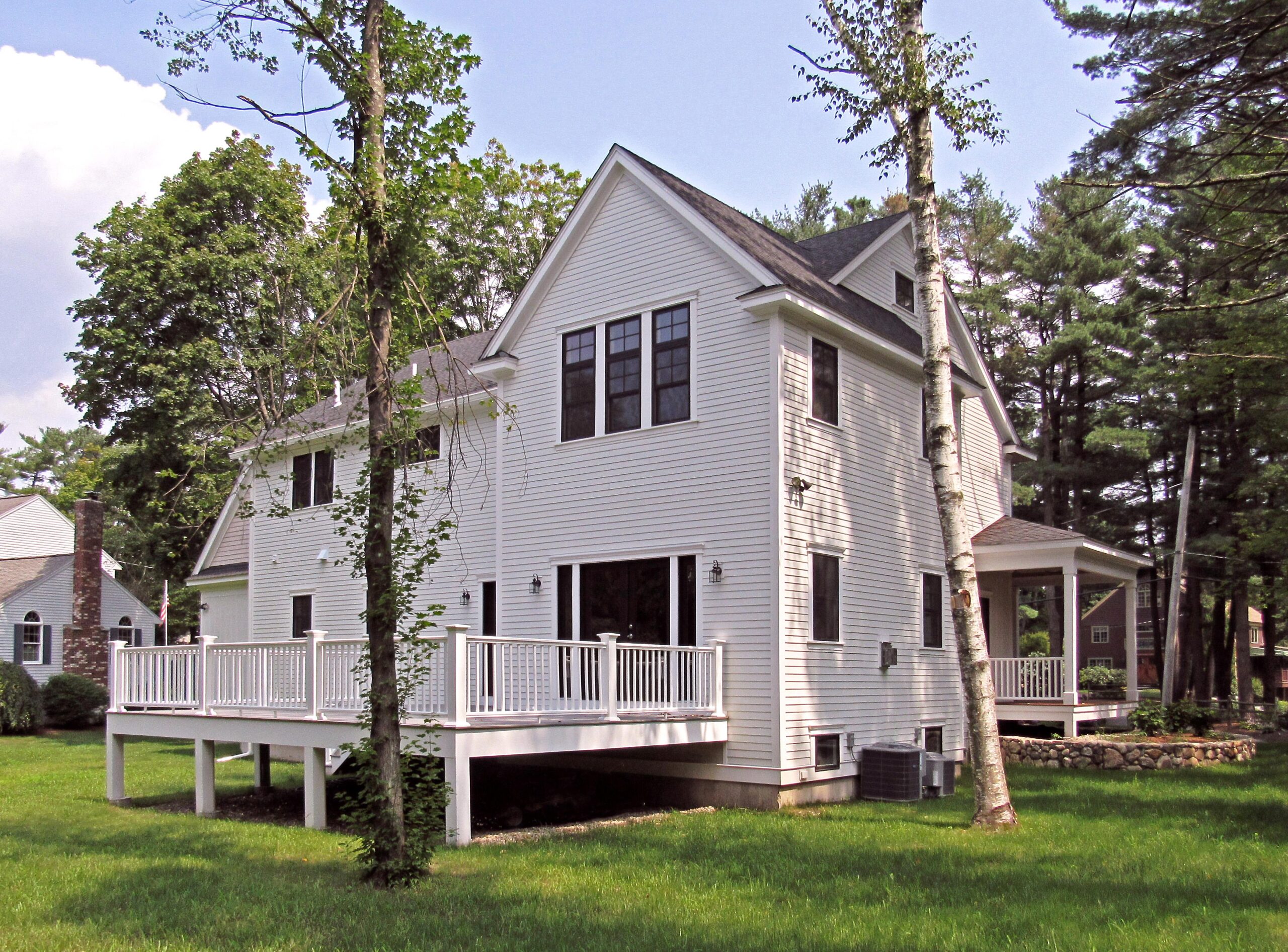 Two-story white house with black-framed windows, elevated deck with white railing, surrounded by green grass and tall trees.