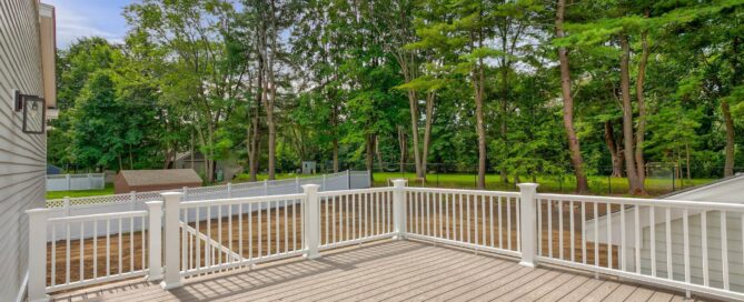 A wooden deck with white railings overlooks a fenced backyard with grass and mature trees under a blue sky.