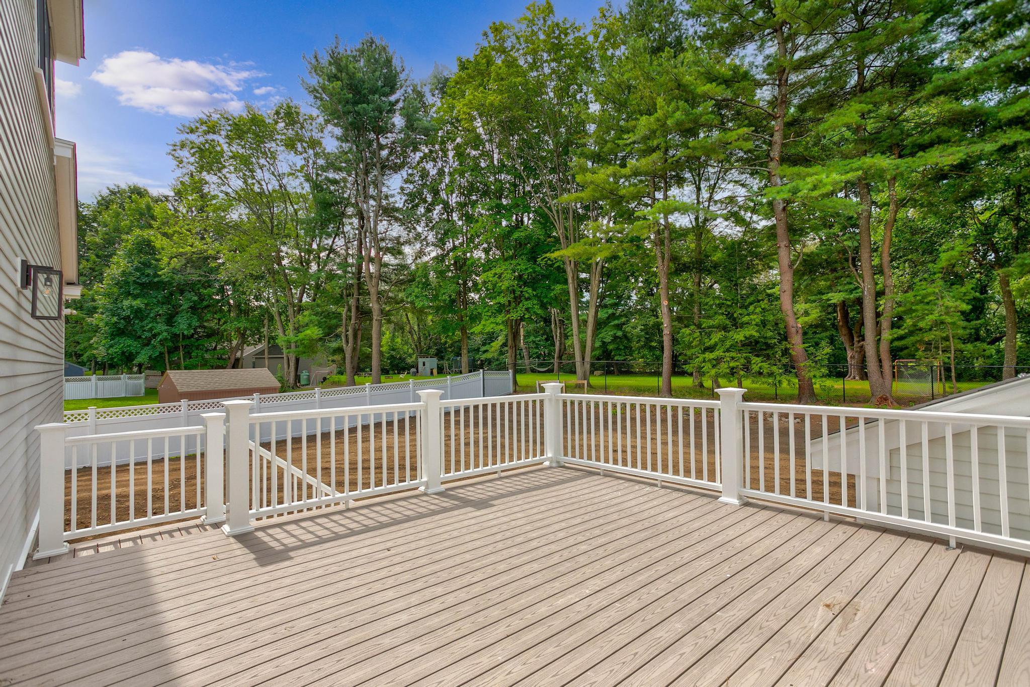 A wooden deck with white railings overlooks a fenced backyard with grass and mature trees under a blue sky.