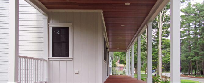 A covered wooden porch with white columns and railings, attached to a white house, overlooks a yard with greenery and trees in the background.