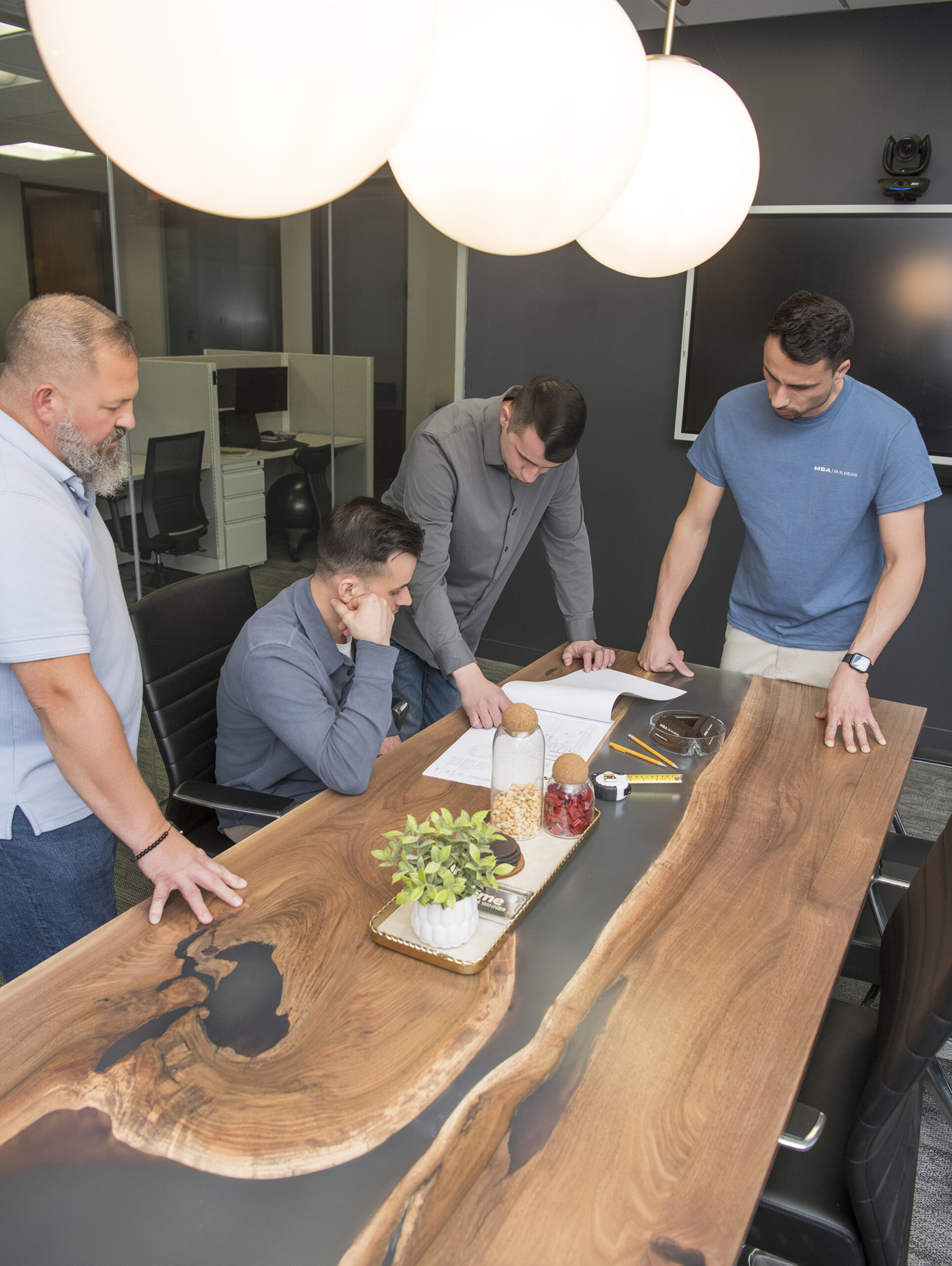 Four men stand and sit around a wooden conference table, reviewing documents related to home design services together in a modern office meeting room. MBA Builders team of experts in home design services