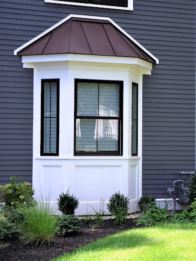 A white bay window with black trim and a brown metal roof enhances the dark gray siding exterior above a landscaped garden bed, showcasing expert exterior home renovation MA window services. A white bay window with black trim and a brown metal roof enhances the dark gray siding exterior above a landscaped garden bed, showcasing expert exterior home renovation MA window services.