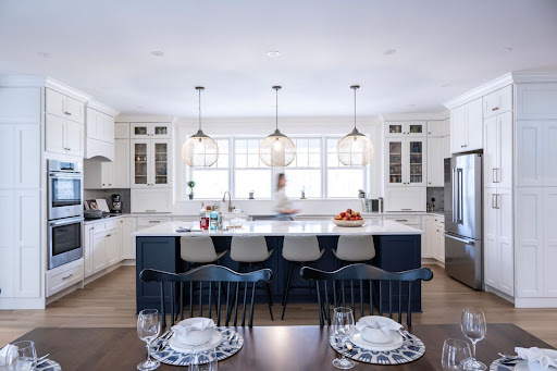 Modern kitchen with white cabinets, a dark blue island, three pendant lights, stainless steel appliances, and a blurred person standing at the sink.