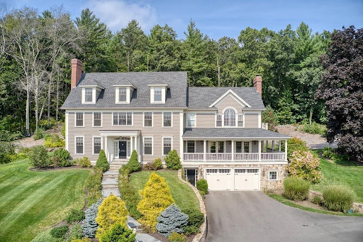 A large two-story house with gray siding, white trim, and a covered front porch, surrounded by landscaped gardens and a driveway leading to a double garage.