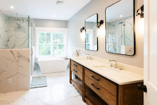 Modern bathroom with double sinks, two mirrors, wooden vanity, wall sconces, a freestanding bathtub, large window, and marble surfaces on the floor and shower walls.