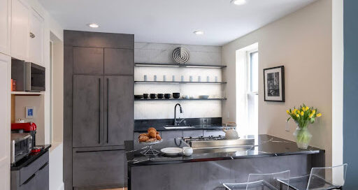 Modern kitchen with dark cabinets, open shelves holding dishes, a black countertop island with pastries and a teapot, two clear chairs, and a vase of yellow tulips—showcasing features that can impact kitchen renovation cost.