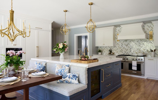 Modern kitchen with white cabinets, a blue island with built-in seating, gold light fixtures, and stainless steel appliances—showcasing style that maximizes value for your kitchen renovation cost. A dining table is set with flowers and tableware.