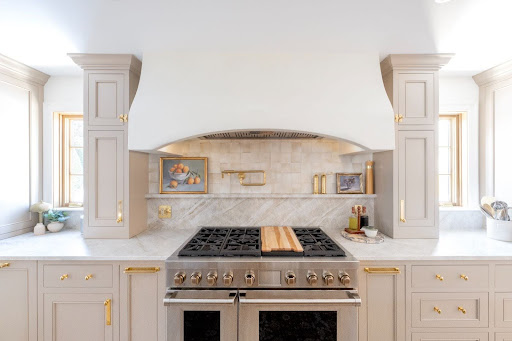 Modern kitchen with a stainless steel gas stove, wooden cutting board, light grey cabinets, marble countertops, and brass fixtures—plus ample natural light. A beautiful result that showcases the value of your kitchen renovation cost.