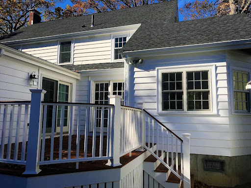 White two-story house with a dark gray roof, multiple windows, and a wooden deck with white railings and stairs leading to the yard. Trees with autumn leaves are in the background.