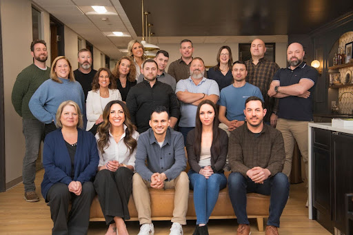 A group of nineteen adults pose indoors, with some seated on a couch and others standing behind them, all facing the camera in a well-lit office setting.