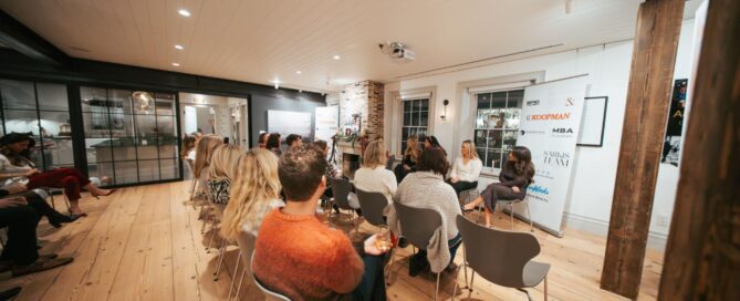 People seated in a modern room listen to two women speaking at the front, next to a sign with logos, during a panel discussion or presentation.