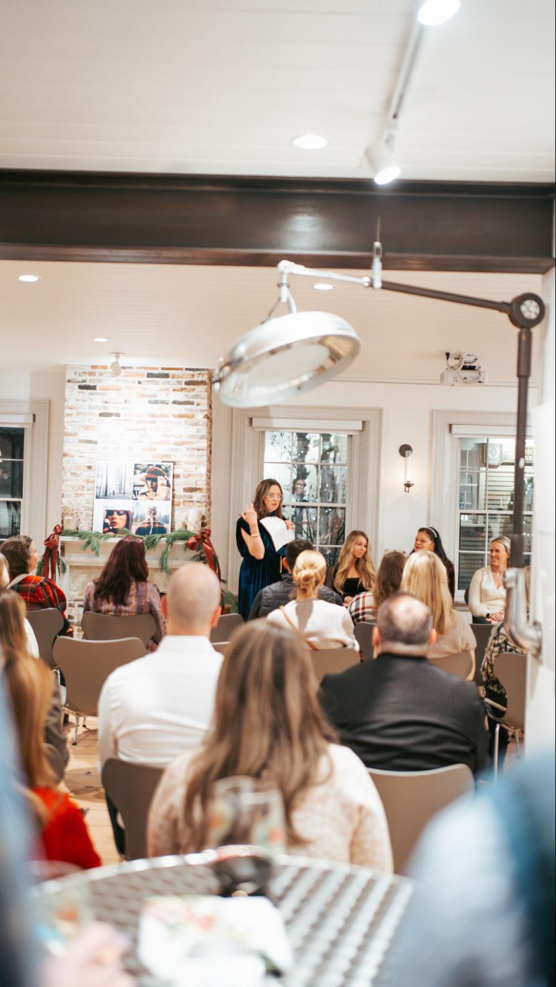 A woman stands and speaks to an audience seated in a brightly lit room with exposed brick and modern decor.