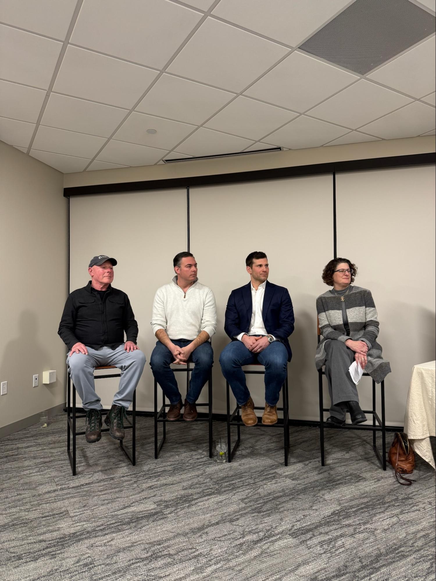 Four people sit on high chairs in a row against a beige wall in a conference room, facing forward, as they discuss ADU Builder Massachusetts projects.