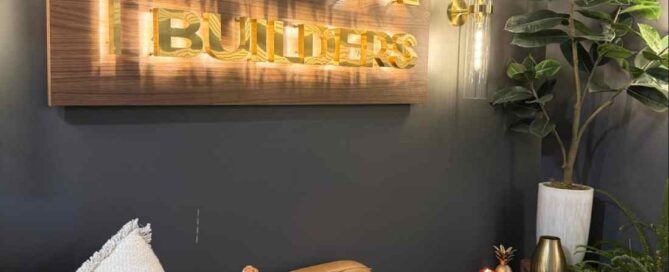A modern office lounge with a brown leather couch, decorative pillows, a small table with books and a candle, a potted plant, and a lit "MBA BUILDERS" wall sign—perfect inspiration for any ADU Builder in Massachusetts.
