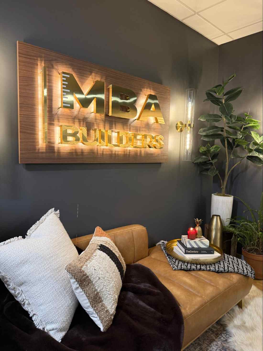 A modern office lounge with a brown leather couch, decorative pillows, a small table with books and a candle, a potted plant, and a lit "MBA BUILDERS" wall sign—perfect inspiration for any ADU Builder in Massachusetts.