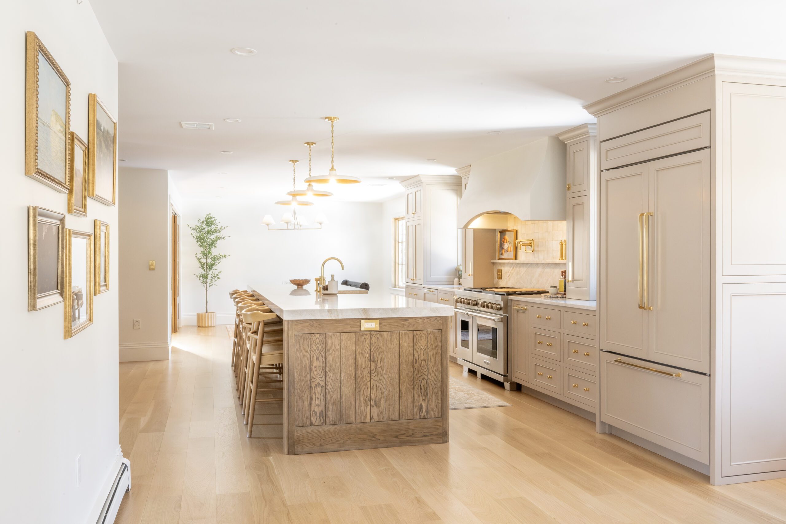 Bright, modern kitchen with light wood floors, a large island with stools, beige cabinetry, stainless steel appliances, gold accents, and framed artwork on the wall.