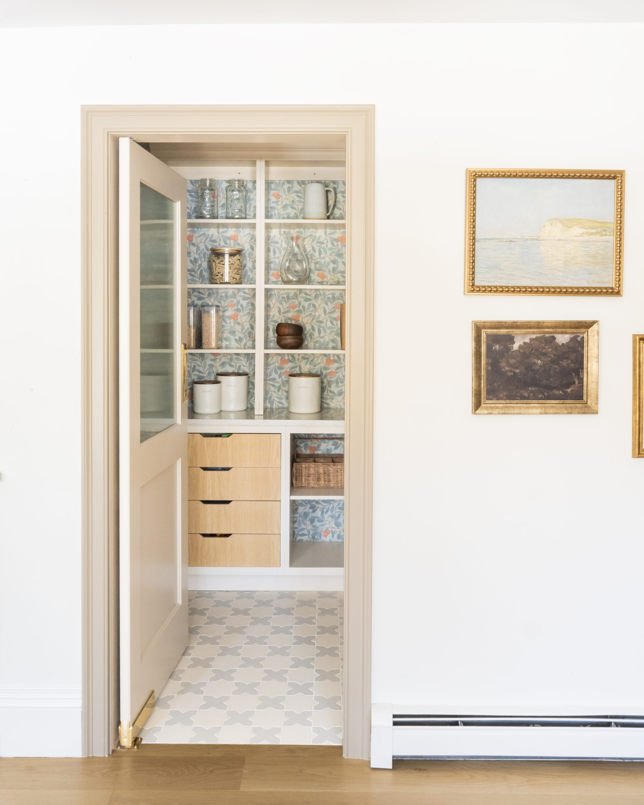 A small pantry with open shelving, canisters, pitchers, and jars, featuring patterned wallpaper and a partially open door. Three framed pictures hang on the adjacent white wall.