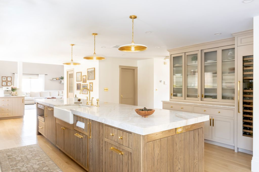 Spacious modern kitchen with a large marble island, wood cabinets, brass fixtures, glass-front storage, and pendant lighting. Adjacent open living area is visible in the background. Spacious modern kitchen with a large marble island, wood cabinets, brass fixtures, glass-front storage, and pendant lighting. Adjacent open living area is visible in the background.
