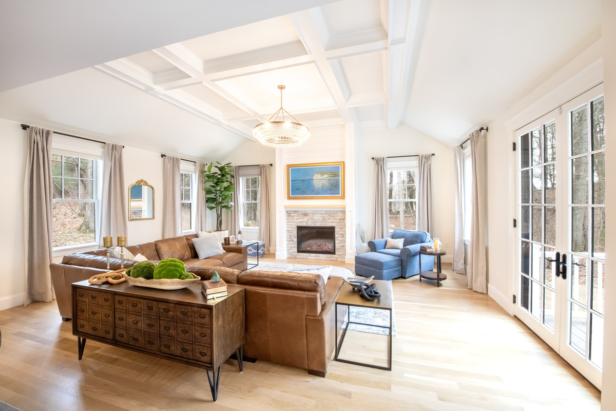 Bright living room with brown leather sectional, blue armchair, wooden floors, fireplace, large windows, and glass doors opening to an outdoor area. Bright living room with brown leather sectional, blue armchair, wooden floors, fireplace, large windows, and glass doors opening to an outdoor area.