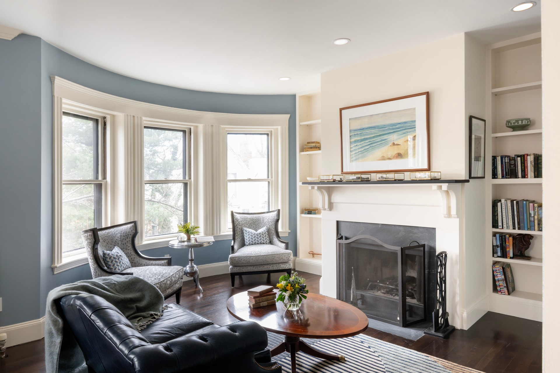 Bright living room with blue walls, a fireplace, bookshelves, two armchairs by bay windows, a dark sofa, and a round wooden coffee table with flowers. Bright living room with blue walls, a fireplace, bookshelves, two armchairs by bay windows, a dark sofa, and a round wooden coffee table with flowers.