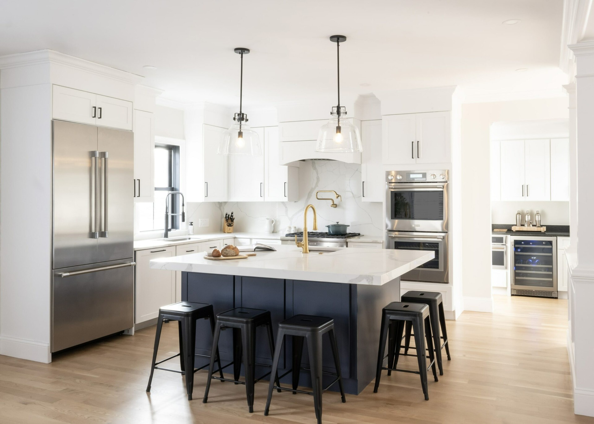 Modern kitchen with white cabinets, stainless steel appliances, a large island with black stools, pendant lights, and wood flooring. Modern kitchen with white cabinets, stainless steel appliances, a large island with black stools, pendant lights, and wood flooring.