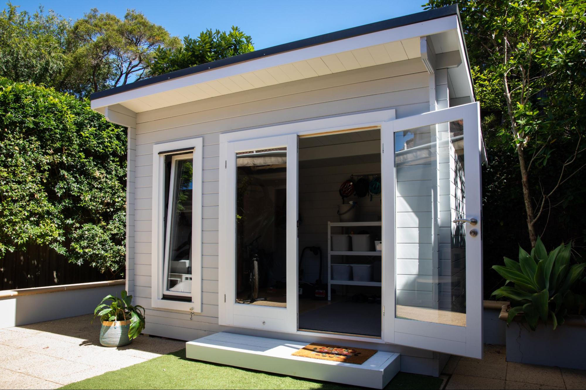 A light grey garden shed with glass double doors, a single tilt-window, and a small step, set in a sunny backyard.