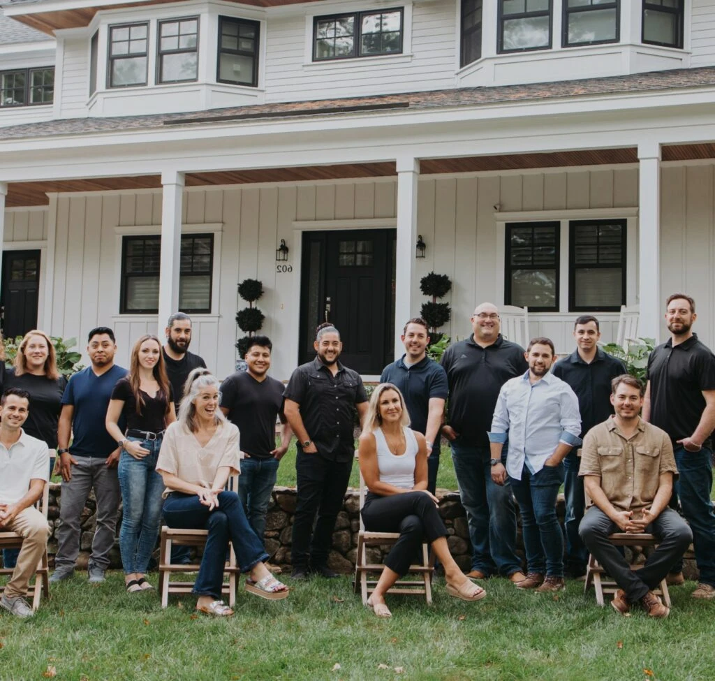 A group of diverse adults smiling, standing, and sitting in front of a two-story building with white siding and black accents, representing their journey in building relationships with MBA Builders. A group of diverse adults smiling, standing, and sitting in front of a two-story building with white siding and black accents, representing their journey in building relationships with MBA Builders.